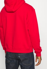 Rotes Hoodie mit geräumiger Kapuze, gerippten Bündchen und Saum. Weicher Stoff mit glatter Textur. Wird mit dunkler Jeans getragen.