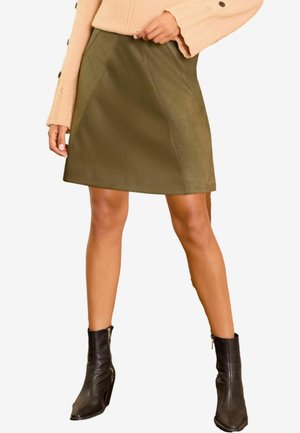 Olive rohe faux nahk A-line seelik sujuva tekstuuriga, keskmise reie pikkusega ja kahe keskkohaga õmblusega, koos mustade pahkluute saapatega.