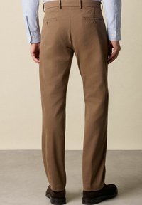 Uomo che indossa pantaloni dritti marroni, scarpe marroni scure e una camicia a righe blu, in piedi su un pavimento grigio contro un muro beige.
