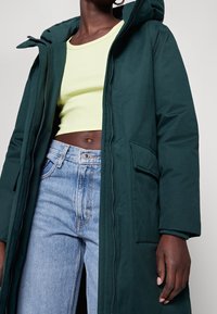 Mørkegrøn, hætteparka med lynlås og frontlommer. Sammen med en lys gul crop top og lyseblå jeans. Glat tekstur.