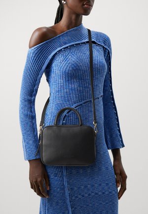 Sort læderhåndtaske med top-håndtag og justerbar rem, med en glat tekstur og minimalistisk design. Bæres tværs over kroppen med en blå ribstrikket kjole.