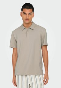 Kortærmet polo t-shirt i lys beige tekstureret materiale, med klassisk krave og diskret knaplukning, parret med stribede bukser.