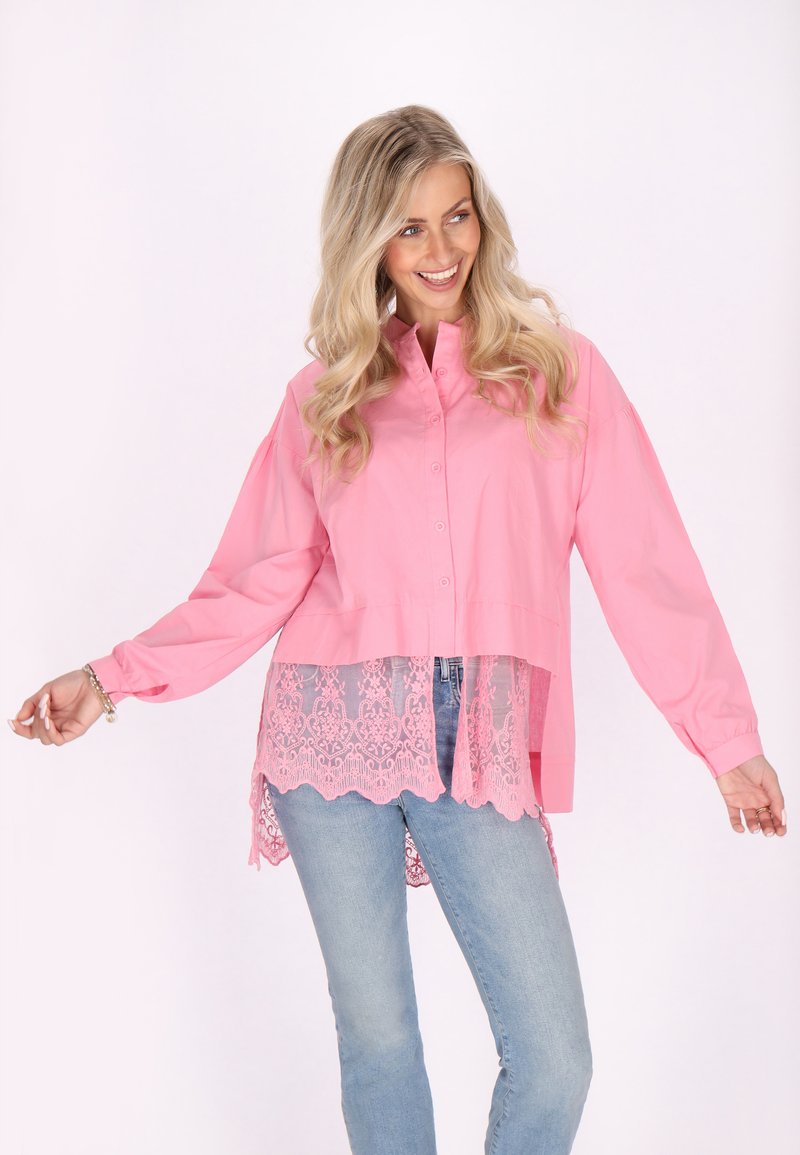 Camicia rosa con bottoni, maniche lunghe, con orlo in pizzo trasparente e bordo decorativo a merletto, abbinata a jeans blu chiaro.
