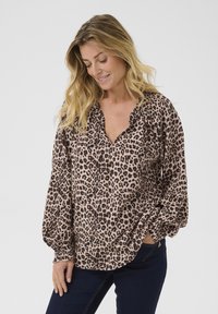 Blouse imprimé léopard en beige clair avec des taches foncées, dotée d'un décolleté en V, de manches froncées et de longues manches amples. Portée avec un jean noir.
