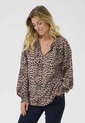 Blouse imprimé léopard en beige clair avec des taches foncées, dotée d'un décolleté en V, de manches froncées et de longues manches amples. Portée avec un jean noir.