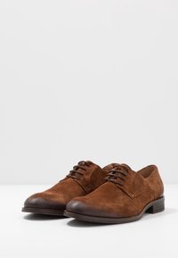 Chaussures oxford en daim marron avec un embout en cuir lisse, présentant un design à lacets à quatre œillets et un petit talon empilé.