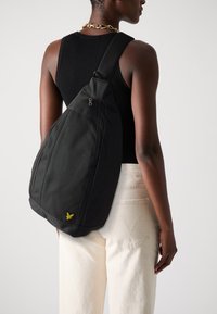 Svart sling ryggsekk med glidelås, laget av slitesterk stoff. Har en gul logo og et stilrent, ergonomisk design. Bæres på ryggen.