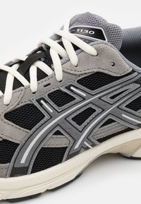 Svart och grå idrottssko med en överdel av meshnät, strukturerade mockadetaljer, reflekterande inslag och krämfärgade snören med en rundad form.