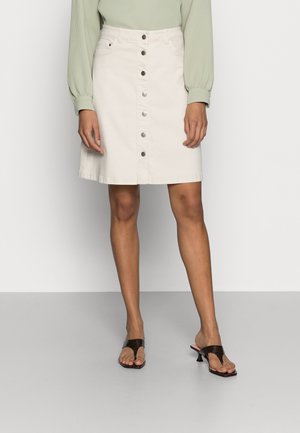 Femme portant une jupe beige boutonnée devant, un haut vert clair à manches longues et des sandales noires à talons, debout sur un sol gris.