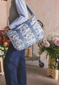 Isik kannab sinimustritatud sinivalget suurt spordikotti, millel on linnavaate disain, seistes lillekorralduste ja ratastega käruga.