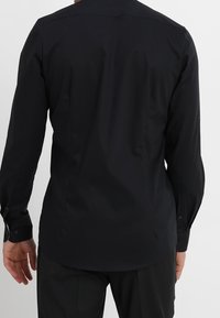 Svart långärmad skjorta med knappkrage, slät tyg, figursydd design och manschetter med en knapp. Detaljerad söm baktill syns.