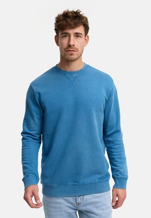 Mężczyzna z krótkimi kręconymi włosami, ubrany w niebieską bluzę z okrągłym dekoltem i jasnoniebieskie dżinsy, stoi na tle jednolitego białego tła.