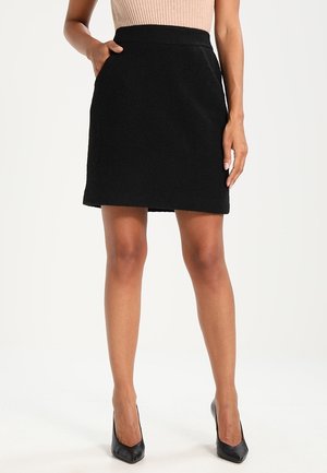 Jupe noire texturée avec une taille cintrée et deux poches latérales, portée avec des talons hauts noirs. Silhouette simple et droite.