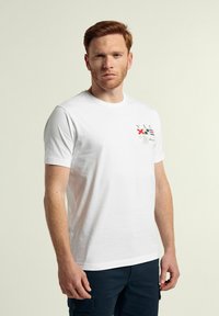 Camiseta de algodón blanca con mangas cortas, cuello redondo, que presenta texto y gráficos multicolores en el lado izquierdo. Textura suave.
