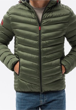 Mężczyzna w oliwkowej pikowanej kurtce puchowej z kapturem, czarnym zamkiem błyskawicznym i rękami w kieszeniach, w połączeniu z niebieskimi dżinsami.