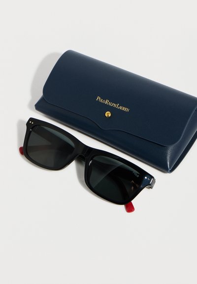 Μαύρα ορθογώνια γυαλιά ηλίου με κόκκινες άκρες και σκούρους φακούς δίπλα σε θήκη Polo Ralph Lauren σε σκούρο μπλε χρώμα με χρυσό κουμπί και λογότυπο.