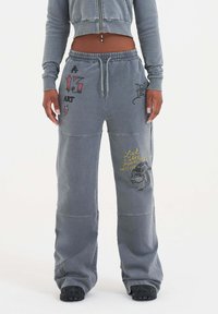 Ljust grå sweatpants med avslappnad passform, som har olika dekorativa tryck, inklusive text och grafiska mönster i kontrasterande färger.