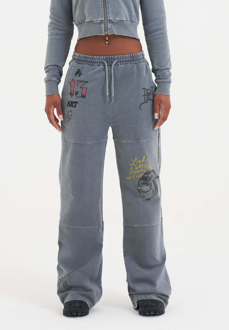 Ljust grå sweatpants med avslappnad passform, som har olika dekorativa tryck, inklusive text och grafiska mönster i kontrasterande färger.