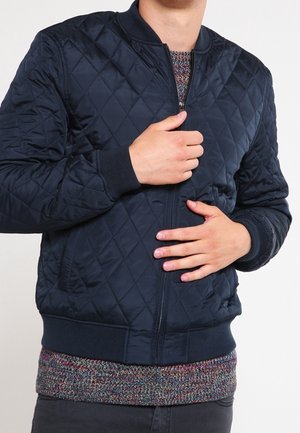 Persoon die een marineblauw gewatteerd bomberjack draagt over een meerkleurige gebreide trui, terwijl hij het jack dichtritst met zichtbaar handen.