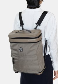 Harmaa kangaskassi, jossa on kova kansi, mustat vetoketjut, säädett�ävät hihnat ja etutasku. Logo näkyvissä edessä.