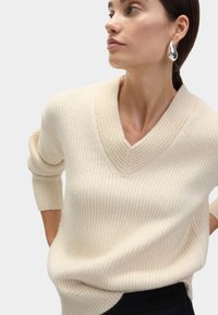 Maglione a coste di colore crema con scollo a V, maniche a tre quarti e vestibilità rilassata, dotato di spacchi laterali per una maggiore comodità.