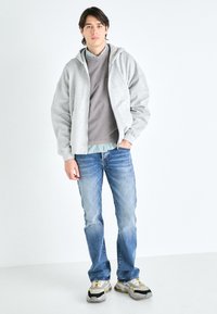 Hallitatud hall kapuutsiga pluus halli triibulise särgi peal, koos helesiniste teksade ja hallide sneakersitega, millel on kollased ja mustad aktsendid.