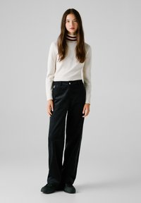Schwarze Cordhosen mit geradem Bein, kombiniert mit einem cremefarbenen Langarmoberteil mit einem dunkelrot gestreiften Ausschnitt. Einfaches Design, minimale Akzente.
