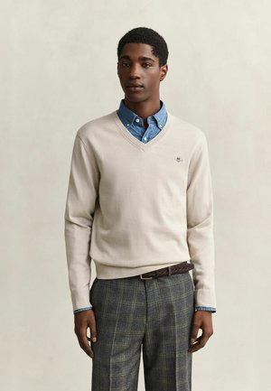 Jonge man die een beige V-hals trui draagt over een blauw overhemd met knopen en grijze geruite broek met een bruine gevlochten riem, staand tegen een effen achtergrond.
