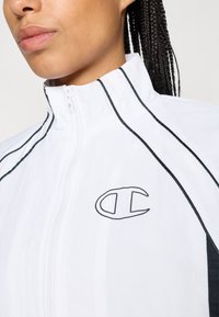 Balts zip-up jaka ar melniem akcentiem un apdari. Viegls audums ar gludu tekstūru, ar logotipu kreisajā pusē.