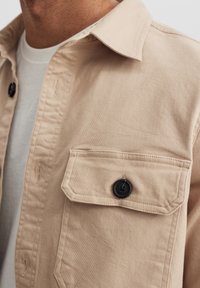 Beige puuvillapaitatakki napituksella, mustat napit, kaksi rintataskua ja klassinen kaulus. Rakenteellinen kangas näkyvillä ompeluilla.