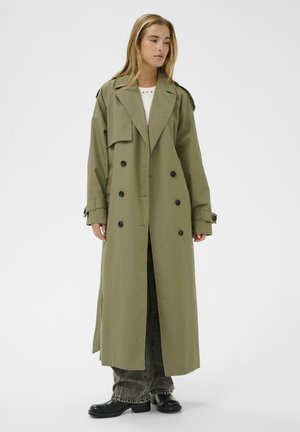 Frau trägt einen olivgrünen langen doppelreihigen Trenchcoat über schwarzen gewaschenen Jeans und schwarzen Stiefeln und steht vor einem einfarbigen Hintergrund.