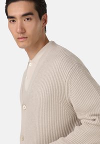 Beżowy sweter dziany z dekoltem w kształcie litery V, zapinany na guziki, o prążkowanej fakturze. Charakteryzuje się gładkim materiałem, delikatnym połyskiem i dopasowanym krojem.