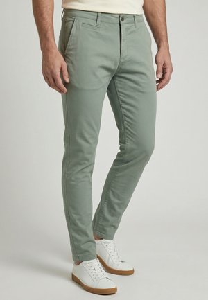 Homme portant un pantalon slim vert clair et des baskets blanches avec semelles en gomme, debout devant un fond clair uni.