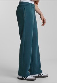 Pantalones de chándal color verde azulado con un corte holgado, confeccionados en una tela suave, que cuentan con bolsillos laterales y una cinturilla acanalada. Combinados con zapatillas blancas y grises.