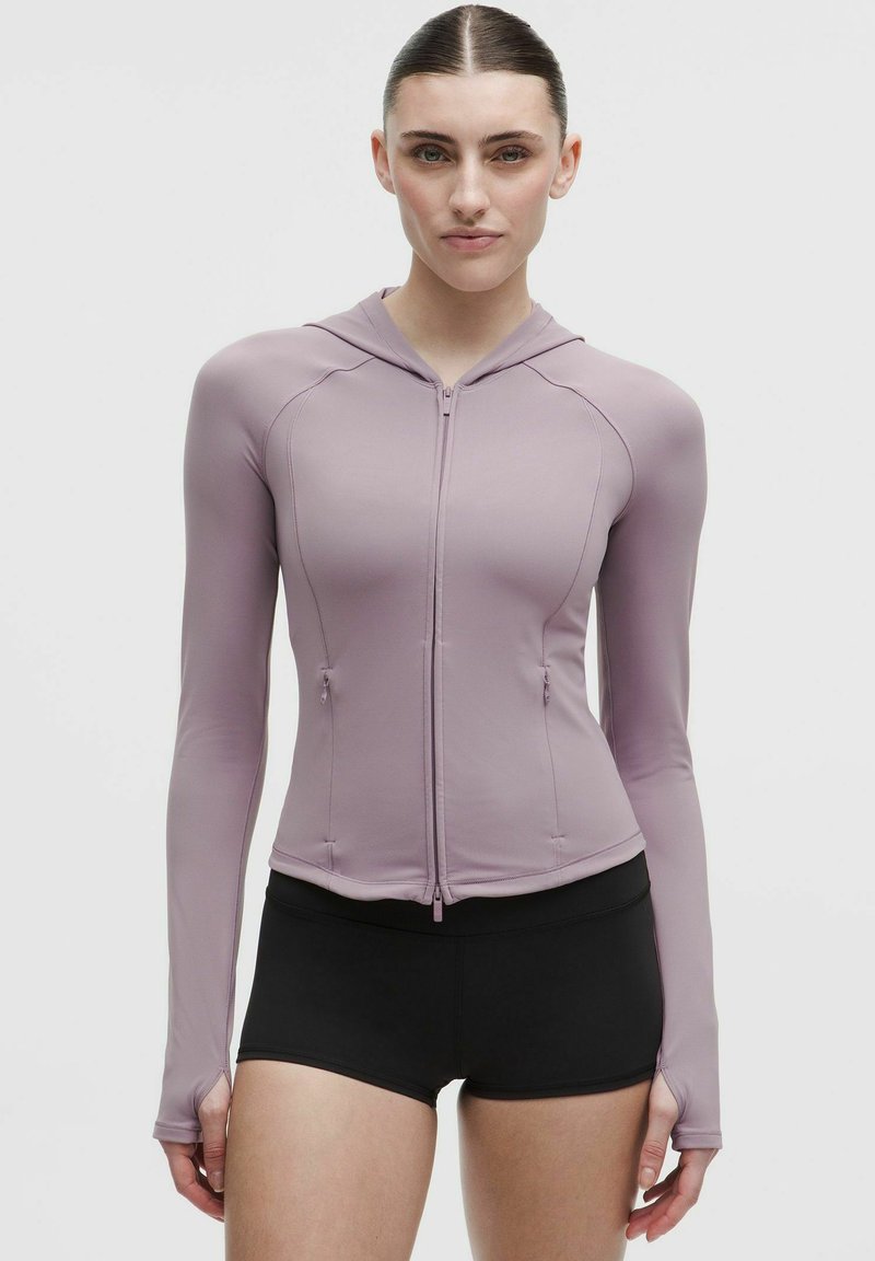 Donna con felpa aderente mauve con zip e fori per i pollici e pantaloncini neri aderenti, in piedi contro uno sfondo chiaro e uniforme.