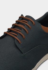 Σκούρο γκρι υφασμάτινο παπούτσι με καφέ στρογγυλά κορδόνια και λευκή σόλα, κοντινή λήψη στα μπροστινά και πλαϊνά στοιχεία.