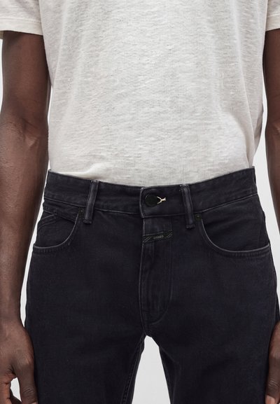 Jean en denim noir avec passants de ceinture et fermeture par bouton, porté par une personne portant une chemise légère texturée à manches courtes.