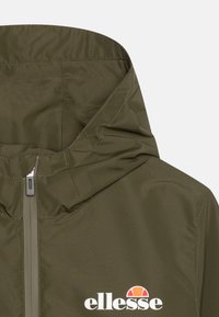 Zielona kurtka z kapturem i przednim zamkiem, z białym logo "ellesse" oraz pomarańczowo-czerwonym półokrągłym emblematem na klatce piersiowej.