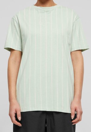 Osoba ubrana w jasnorozieloną koszulkę z krótkim rękawem z cienkimi białymi pionowymi paskami oraz czarne spodnie na neutralnym tle.