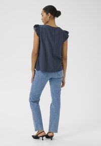Blouse bleu marine à manches courtes volantées, associée à un jean droit bleu clair et des mules noires à talons. Texture de tissu lisse.