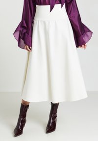 Jupe évasée blanche taille haute, fabriquée en tissu lisse. Associée à un chemisier transparent violet profond et à des bottines brillantes rouge foncé.
