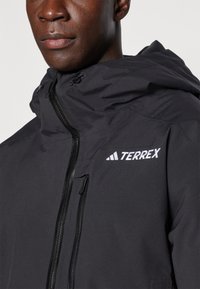 Giacca nera con cappuccio realizzata in materiale testurizzato, dotata di zip frontale e logo bianco "TERREX" sul petto. Design leggero.