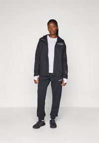 Schwarze Kapuzenjacke mit einem weißen Logo, kombiniert mit einem weißen Hemd und dunkelgrauen Hosen, mit einem lockeren Schnitt und elastischen Bündchen.