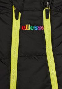 Svart puffjacka med klara gula dragkedjor, med flerfärgad broderad "ellesse"-logotyp på framsidan. Texturerad tyg.