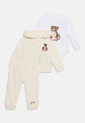 Ensemble bébé trois pièces couleur crème avec veste à capuche, pantalon et body blanc, tous ornés d'un motif ours en peluche et d'un texte rouge "guess".
