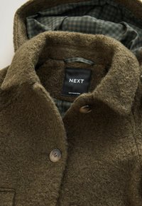 Olivengrøn, struktureret uldfrakke med knaplukning og foret med ternet stof. Har et mærket sort label med "NEXT."