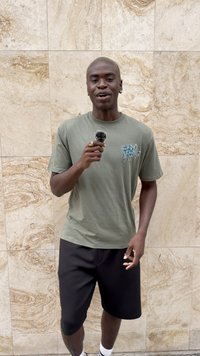 Grünes Baumwoll-T-Shirt mit blauem Grafikdruck, kombiniert mit schwarzen Shorts. Hält ein Mikrofon mit einem pelzigen Windschutz, steht auf einer strukturierten Oberfläche.