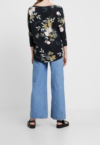 Top floreale nero con maniche a tre quarti, decorato con fiori rosa e dorati, abbinato a jeans a gamba larga blu chiaro e sandali neri.