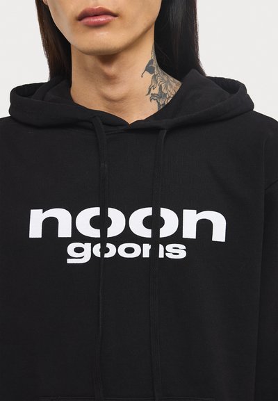 Schwarze Kapuzenjacke aus weichem Stoff mit einem auffälligen weißen Logo, das "noon goons" liest. Sie verfügt über eine Kordel und eine Kängurutasche.