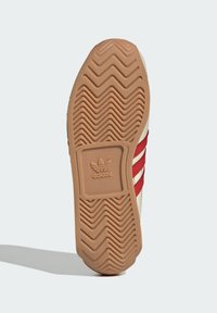 Adidas sneaker sole in gum-colored rubber, featuring a zigzag pattern, with a textured tan base and three red stripes on the side.